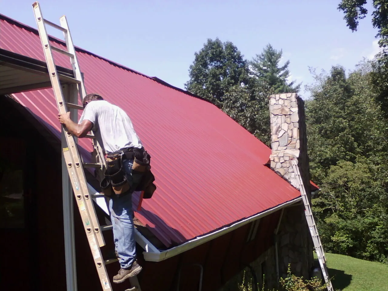 Roofer on a ladder beside a red metal roof and stone chimney — Soffit & Fascia Repair by Roofing Brothers in Slaterville Springs